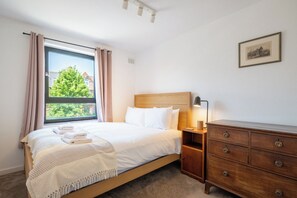 Ein ordentlich bezogenes Bett mit weißen Leinen und einem Kopfteil, ein Nachttisch mit Lampe, eine Holzkommode mit Schubladen und ein Fenster mit Vorhängen.