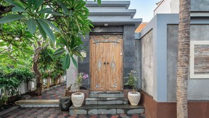 A wooden door with a decorative pattern, a tiled pathway leading to the entrance, and two potted plants on the steps. - Q~la Villas (Legian)