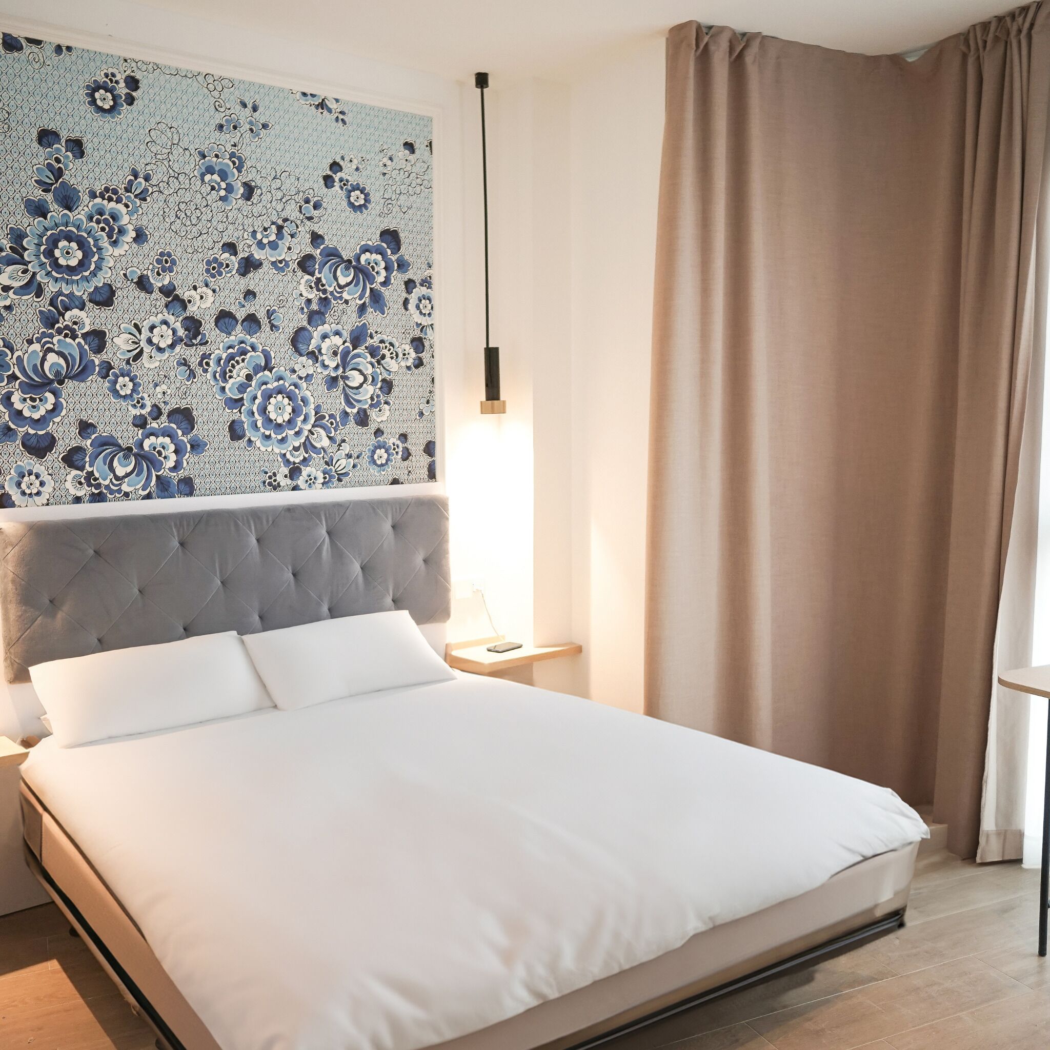 Ein ordentlich bezogenes Bett mit grauem Kopfteil, eine Wand mit Blumenmuster, eine Nachttischlampe und ein Fenster mit Vorhängen.