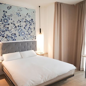 Ein ordentlich bezogenes Bett mit grauem Kopfteil, eine Wand mit Blumenmuster, eine Nachttischlampe und ein Fenster mit Vorhängen.