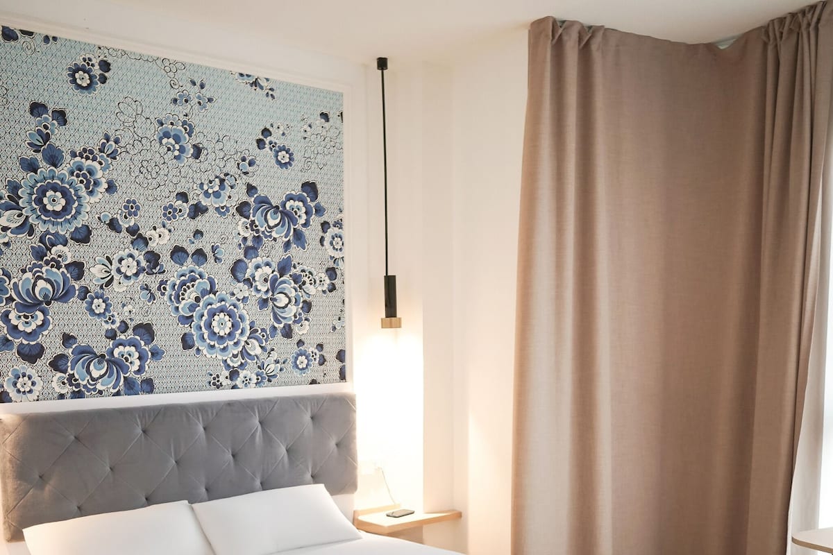 Ein ordentlich bezogenes Bett mit grauem Kopfteil, eine Wand mit Blumenmuster, eine Nachttischlampe und ein Fenster mit Vorhängen.