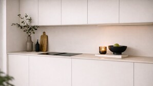 Una cocina moderna con armarios blancos, encimera negra, un florero con hojas verdes, tabla de cortar de madera, una vela y un bol con fruta.