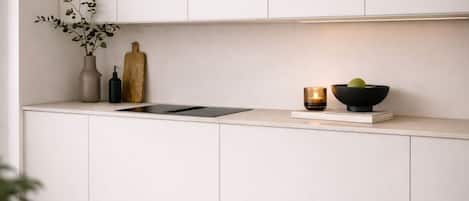 Una cocina moderna con armarios blancos, encimera negra, un florero con hojas verdes, tabla de cortar de madera, una vela y un bol con fruta.