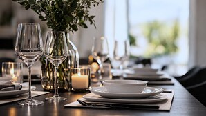 Una mesa de comedor dispuesta con copas de vino, platos y velas, con un florero de follaje al fondo.