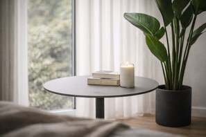 A round table with a candle and books, a potted plant, and a window with blinds.