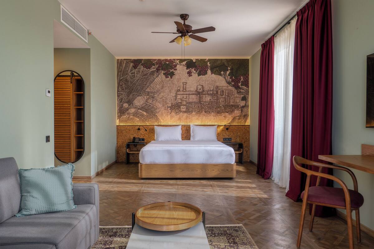 Un dormitorio con una cama, un ventilador de techo, una mesita de noche de madera, una silla, un sofá y un mural en la pared.