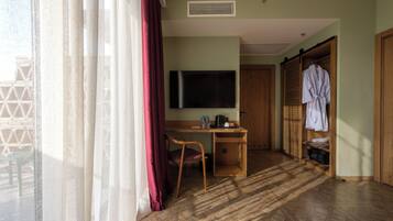 A hotel room with a wooden desk, a television, a robe hanging, and a view through a window with curtains.