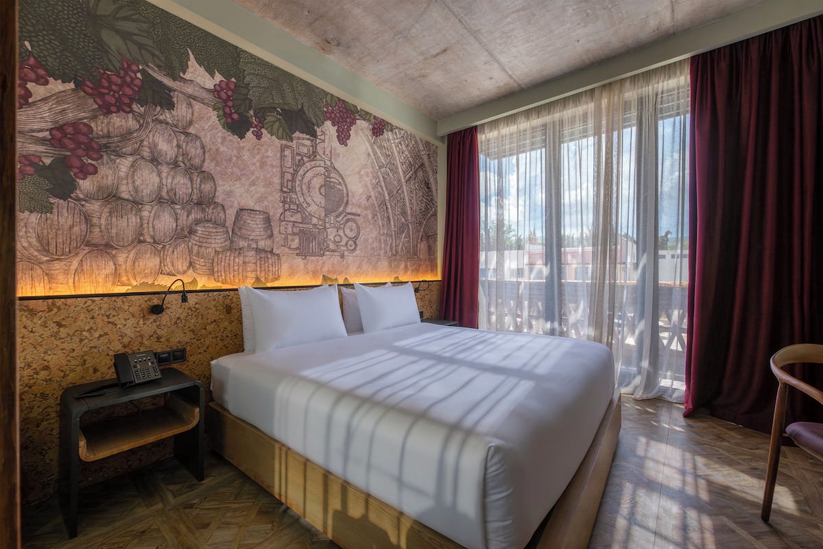 Habitación de hotel con una cama grande, una mesita de noche con teléfono, un mural en la pared y vistas a través de cortinas translúcidas.