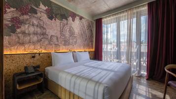 A hotel room with a large bed, a nightstand with a phone, a wall mural, and a view through sheer curtains.