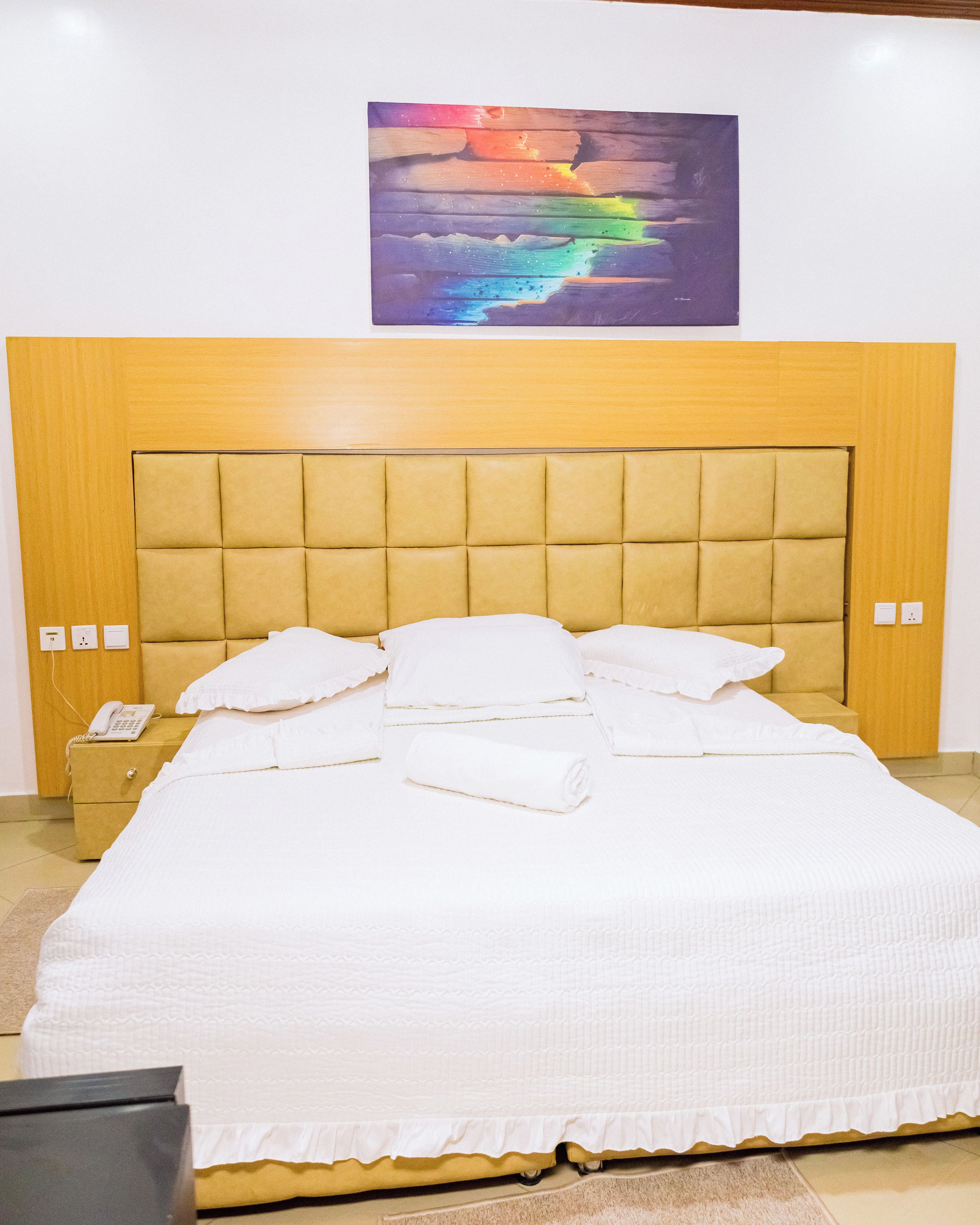 A neatly made bed with a headboard, a bedside phone, and a painting above it.