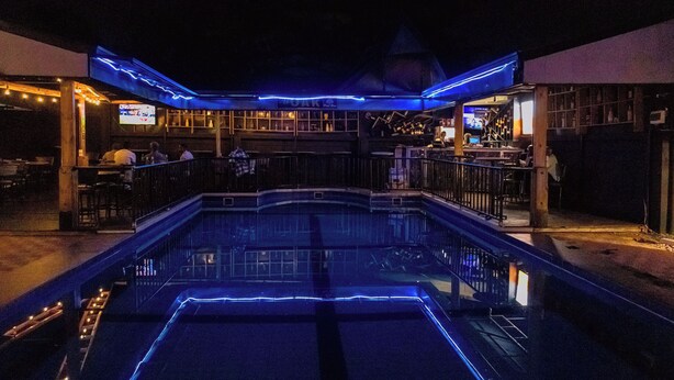 A pool area with a bar and seating, surrounded by a building with a distinctive roof structure. - Poolside Viewbguesthouse (Kigali)