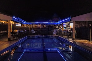 A pool area with a bar and seating, surrounded by a building with a distinctive roof structure. - Poolside Viewbguesthouse (Kigali)