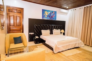 A bedroom with a bed, a chair, a bedside table, a painting on the wall, and a wooden door. - Poolside Viewbguesthouse (Kigali)