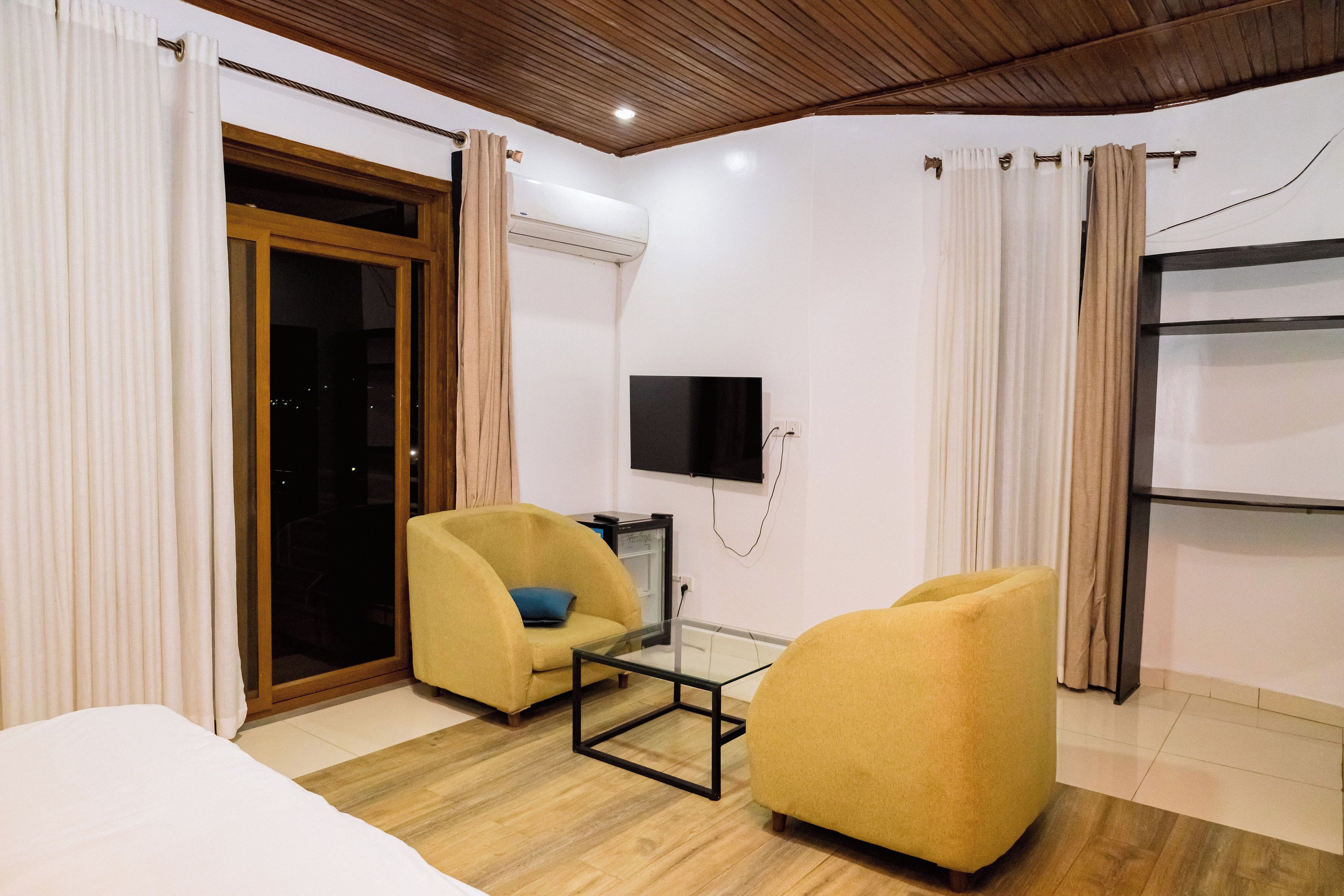 A hotel room with a bed, two yellow armchairs, a glass coffee table, a TV mounted on the wall, and a sliding glass door leading to an outdoor area.