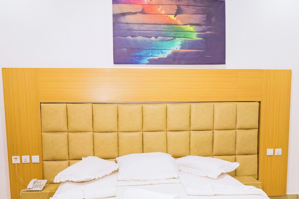 A neatly made bed with a headboard, a bedside phone, and a painting above it.