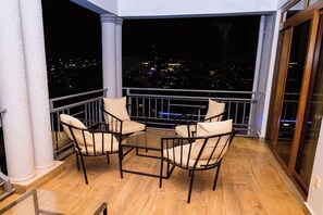 A balcony with a glass table and four chairs, overlooking a cityscape at night. - Poolside Viewbguesthouse (Kigali)
