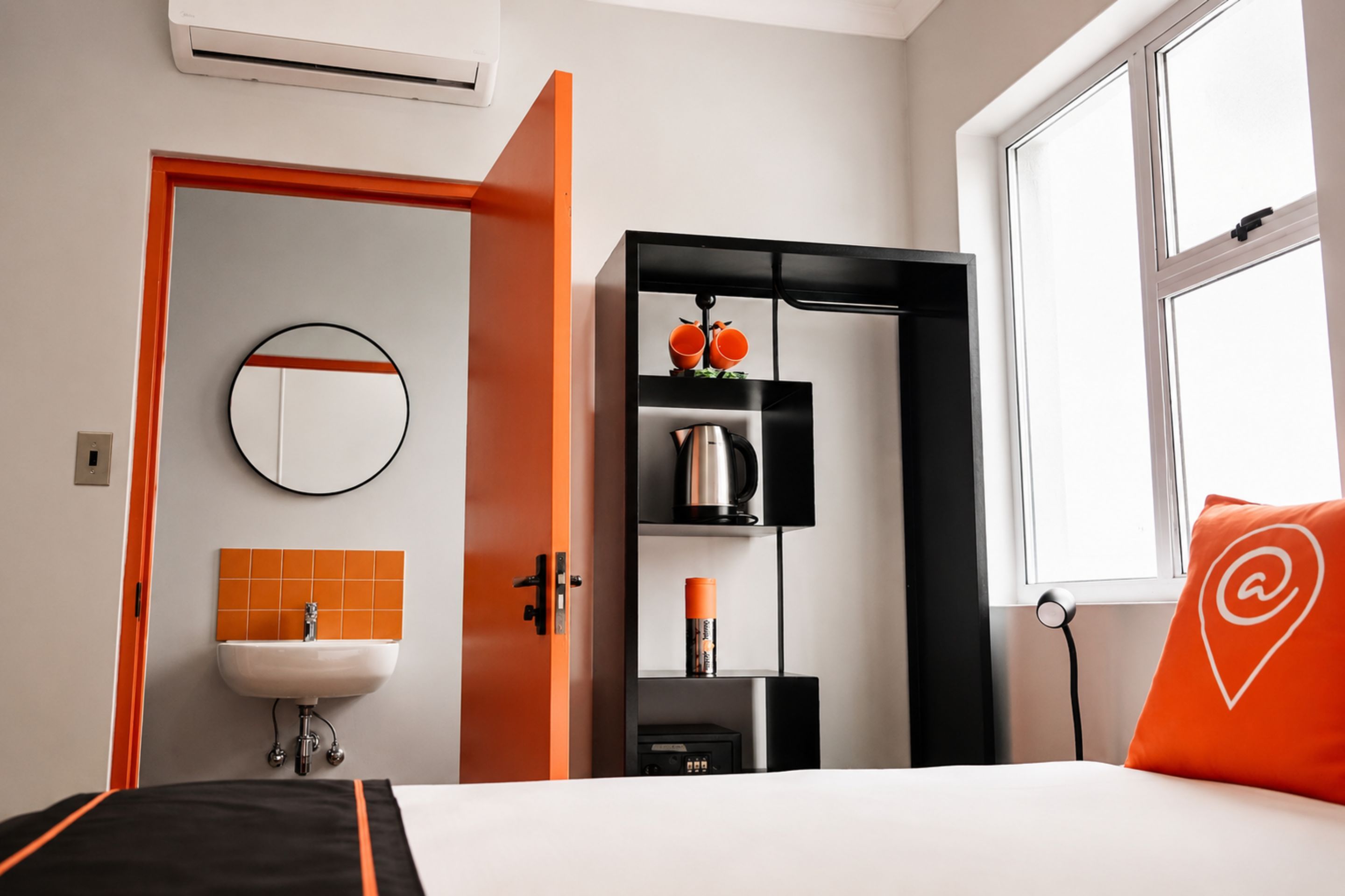 Hotelkamer met een bed, een ronde spiegel, een wastafel, een plank met een waterkoker en een raam met een kussen.