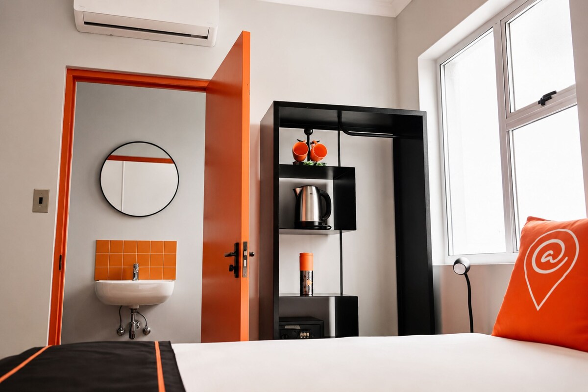 Habitació d'hotel amb un llit, un mirall rodó, un lavabo, una estant amb una tetera i una finestra amb un coixí.