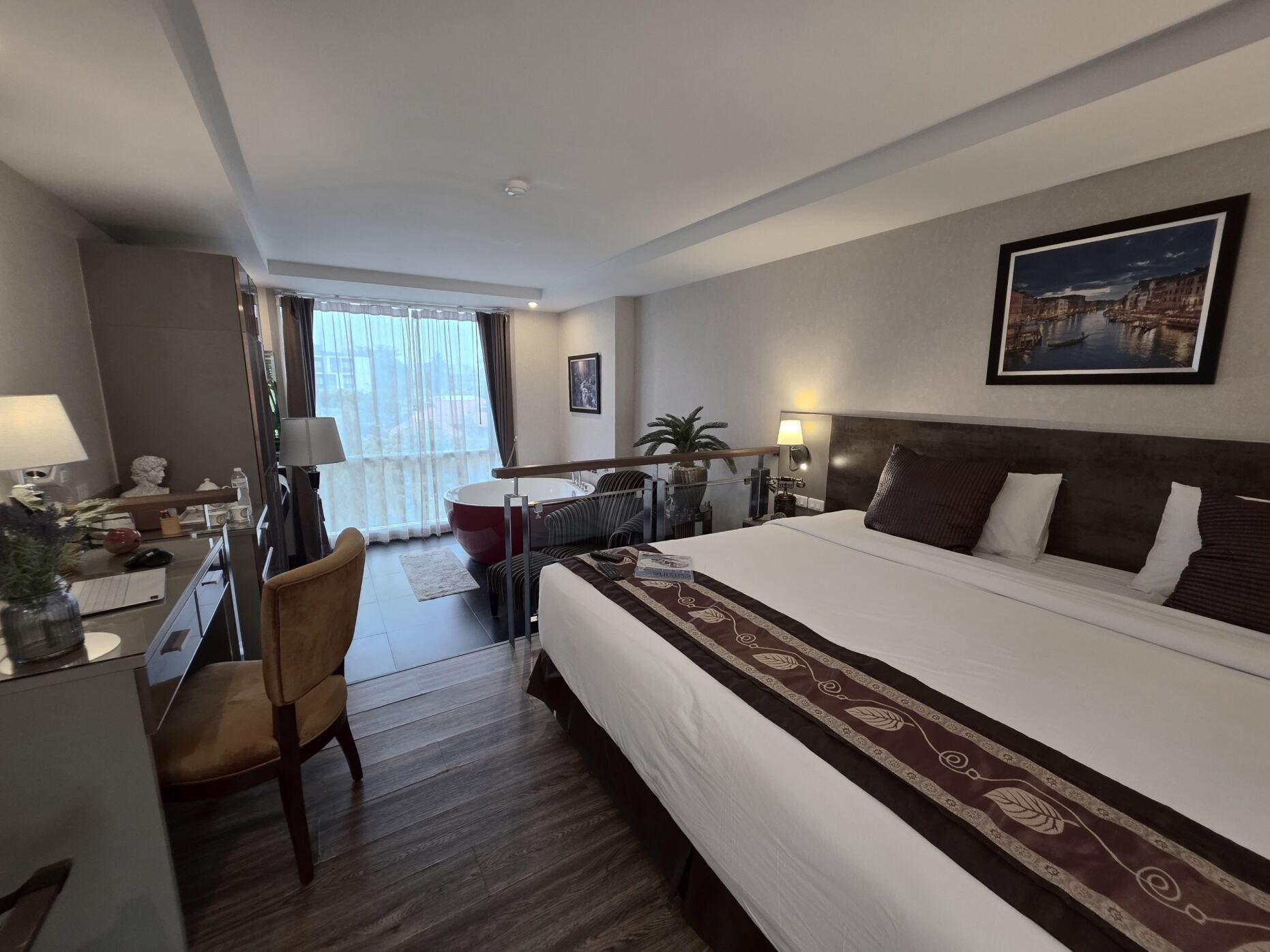 Habitación de hotel con una cama grande, un escritorio con silla, una ventana con cortinas, un cuadro en la pared y una planta en maceta.