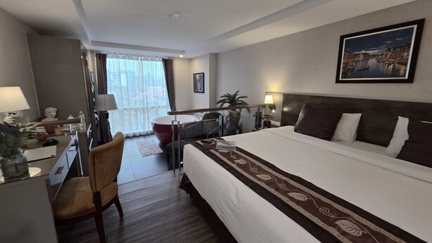 A hotel room with a large bed, a desk with a chair, a window with curtains, a painting on the wall, and a potted plant. - Le Cannes, A Recall Luxe Hotel, Chiang Mai (Chiang Mai)