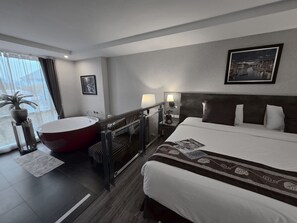 Habitación de hotel con una cama grande, una bañera independiente, una mesita de noche con lámpara, un cuadro en la pared y una planta en maceta cerca de la ventana.