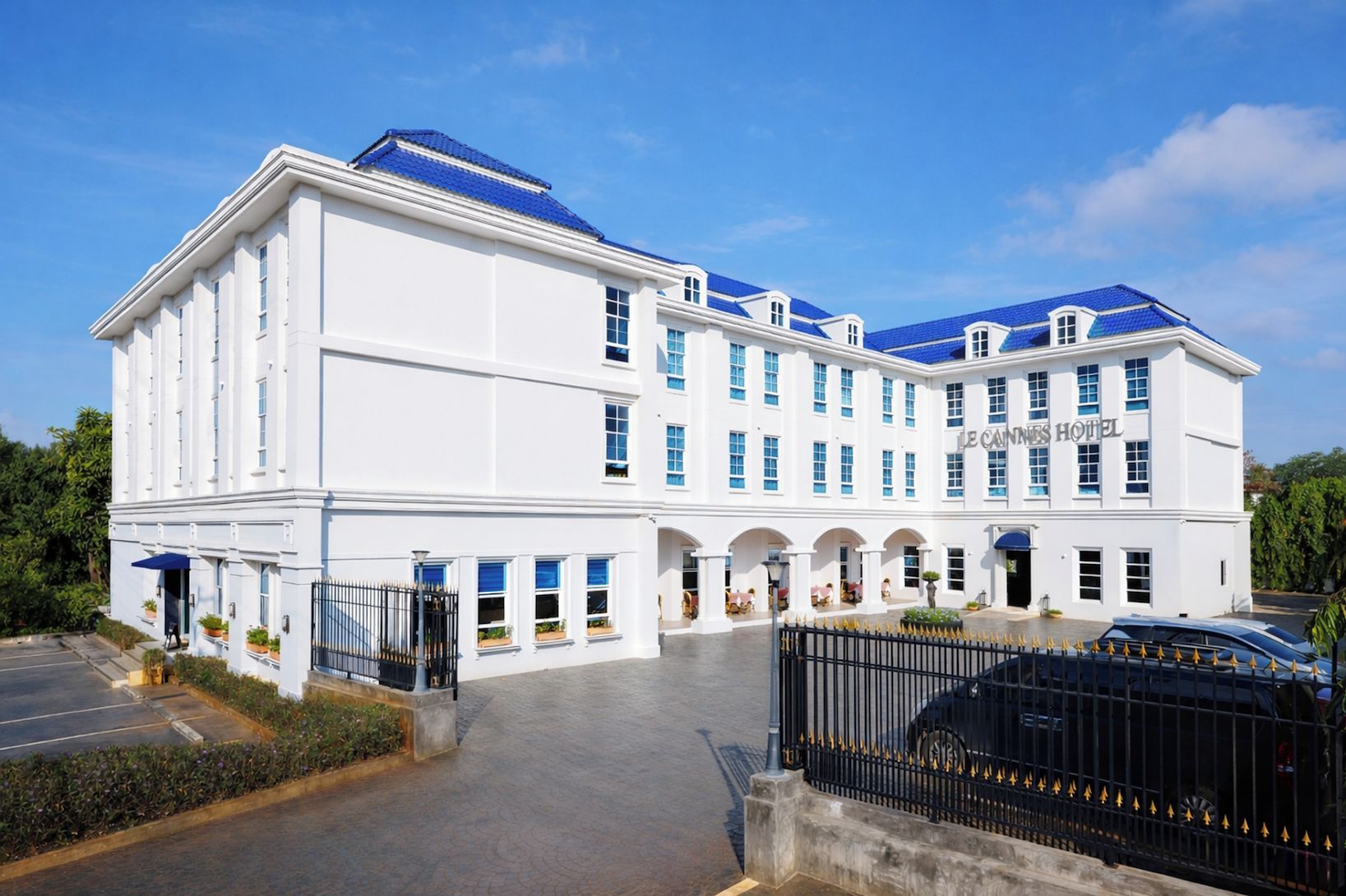 A large, white building with a blue roof, labeled 'Le Caux Hotel,' featuring a spacious entrance and a parking area.