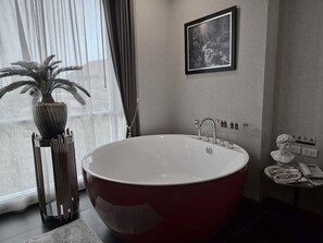 Un baño moderno con una bañera independiente grande, una planta en maceta, una fotografía enmarcada en la pared y una mesita con un teléfono y una revista.