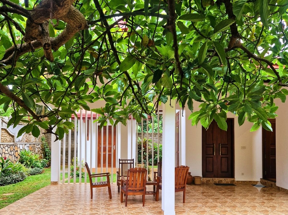 Un'area relax all'aperto coperta, con sedie e un tavolo, circondata da un giardino e un edificio con pavimento piastrellato.