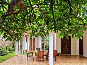 A covered outdoor seating area with chairs and a table, surrounded by a garden and a building with a tiled floor. - Blue Lotus Yoga House (Hikkaduwa)
