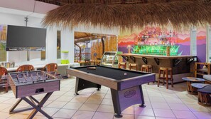 Ein Billardtisch, ein Kicker, eine Bar und Sitzgelegenheiten in einem Raum mit Strohdach und farbenfrohem Wandgemälde.