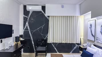 A hotel room with a bed, a desk with a computer, a chair, a TV, and a wall-mounted air conditioner.