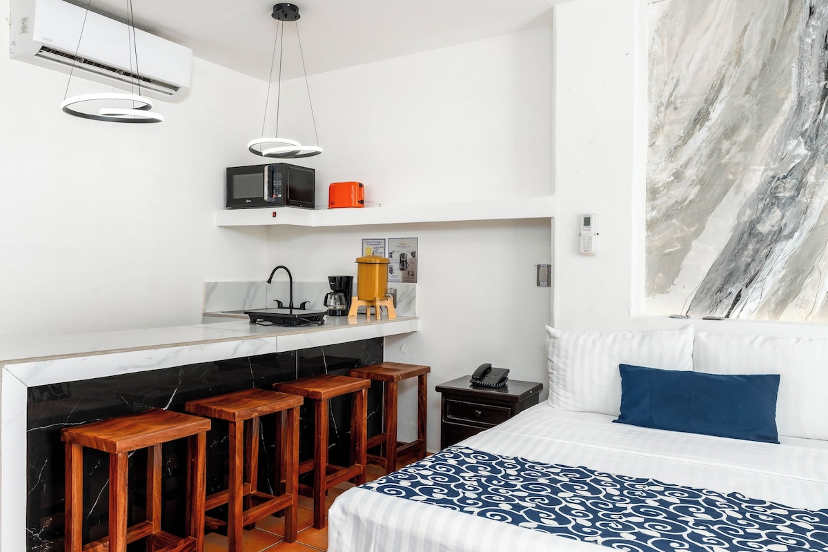 A compact hotel room with a kitchenette, a bed with a patterned bedspread, a bar stool area, and a large abstract wall art piece.