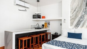 Habitación de hotel compacta con kitchenette, una cama con colcha estampada, zona de taburete de bar y una gran obra de arte abstracto en la pared.
