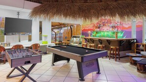 Mesa de billar, mesa de futbolín, zona de bar y asientos en una habitación con techo de paja y mural colorido.