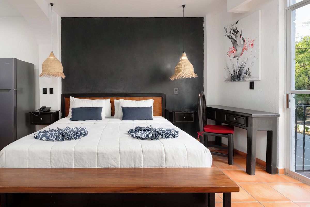 A modern hotel room with a large bed, two bedside tables, a desk, and a kitchenette.