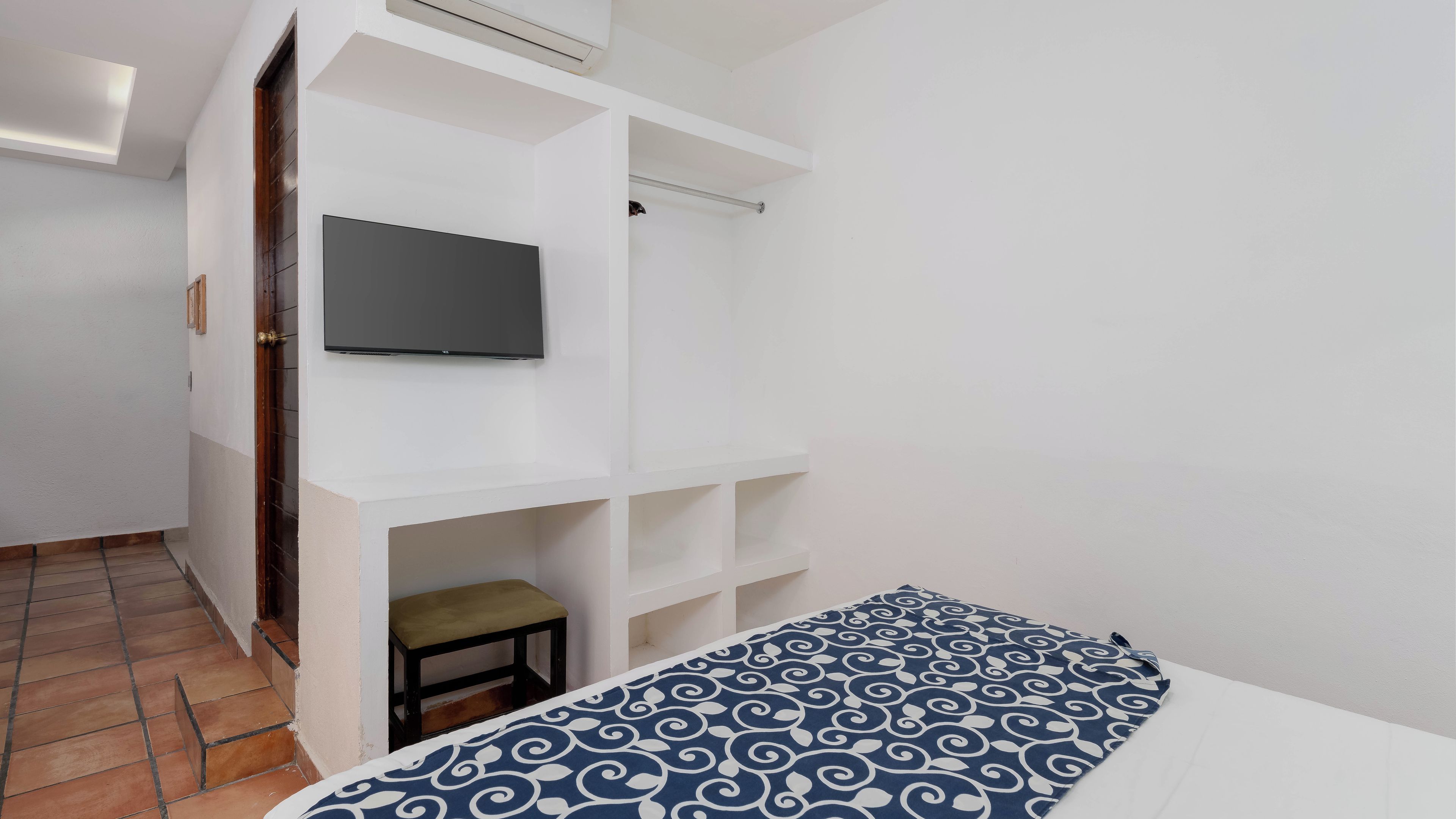 A bedroom with a bed, a TV mounted on a white wall, and a small stool.