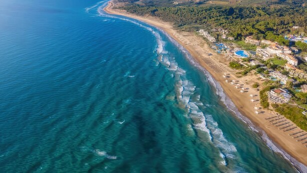 Vista aérea de una zona costera con playa, aguas azules cristalinas y un pequeño pueblo con edificios y vegetación.