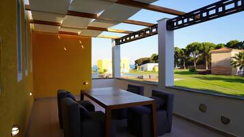 Un patio moderno al aire libre con una mesa y sillas de madera, grandes ventanales que ofrecen vistas a un paisaje verde y edificios.