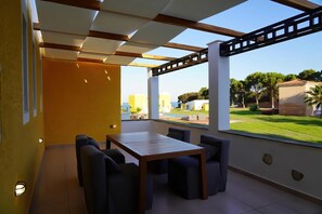 Un patio moderno al aire libre con una mesa y sillas de madera, grandes ventanales que ofrecen vistas a un paisaje verde y edificios.
