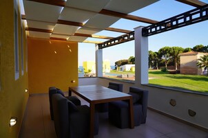 Un patio moderno al aire libre con una mesa y sillas de madera, grandes ventanales que ofrecen vistas a un paisaje verde y edificios.