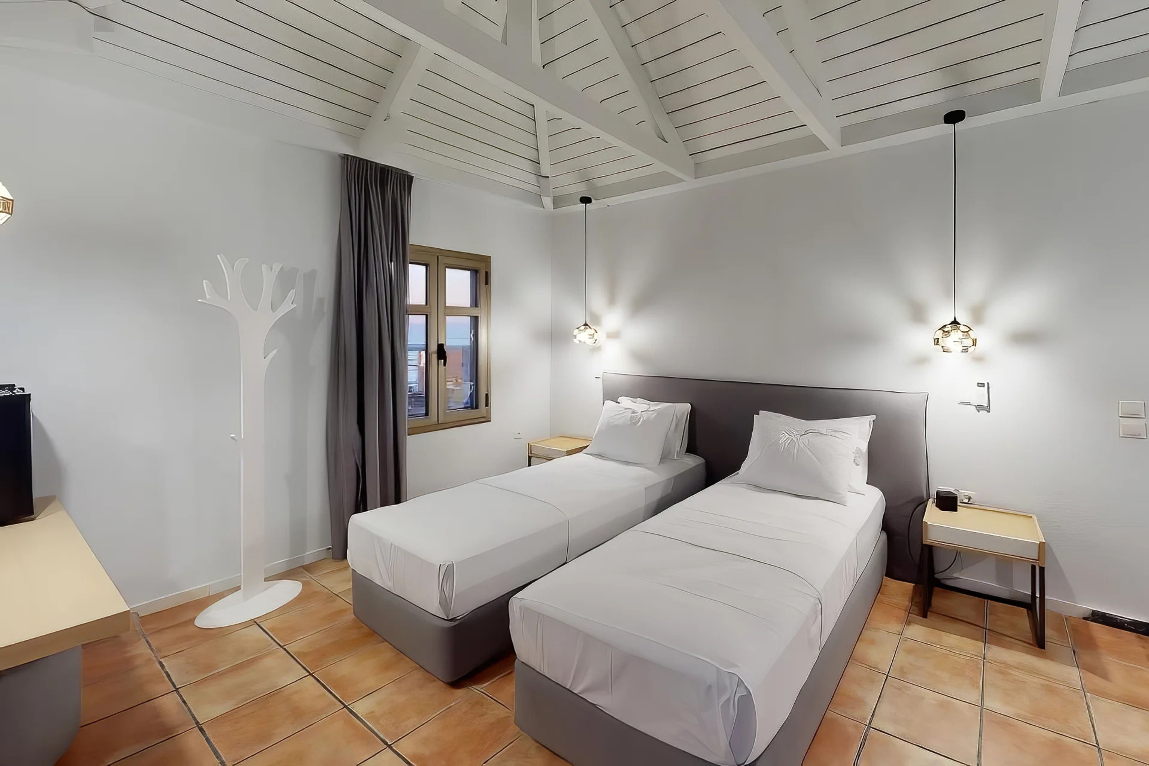 Habitación de hotel con dos camas individuales, un escritorio, una lámpara de pie y un ventanal con cortinas.