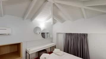 Una habitación de hotel con techo inclinado, una cama, un escritorio con silla, un espejo y una ventana con cortinas.