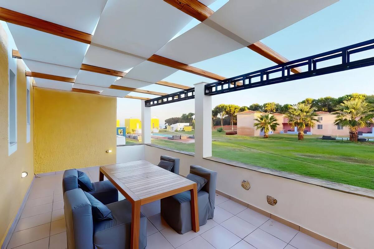A covered outdoor seating area with a table and chairs, large windows with a view of a green lawn and buildings.