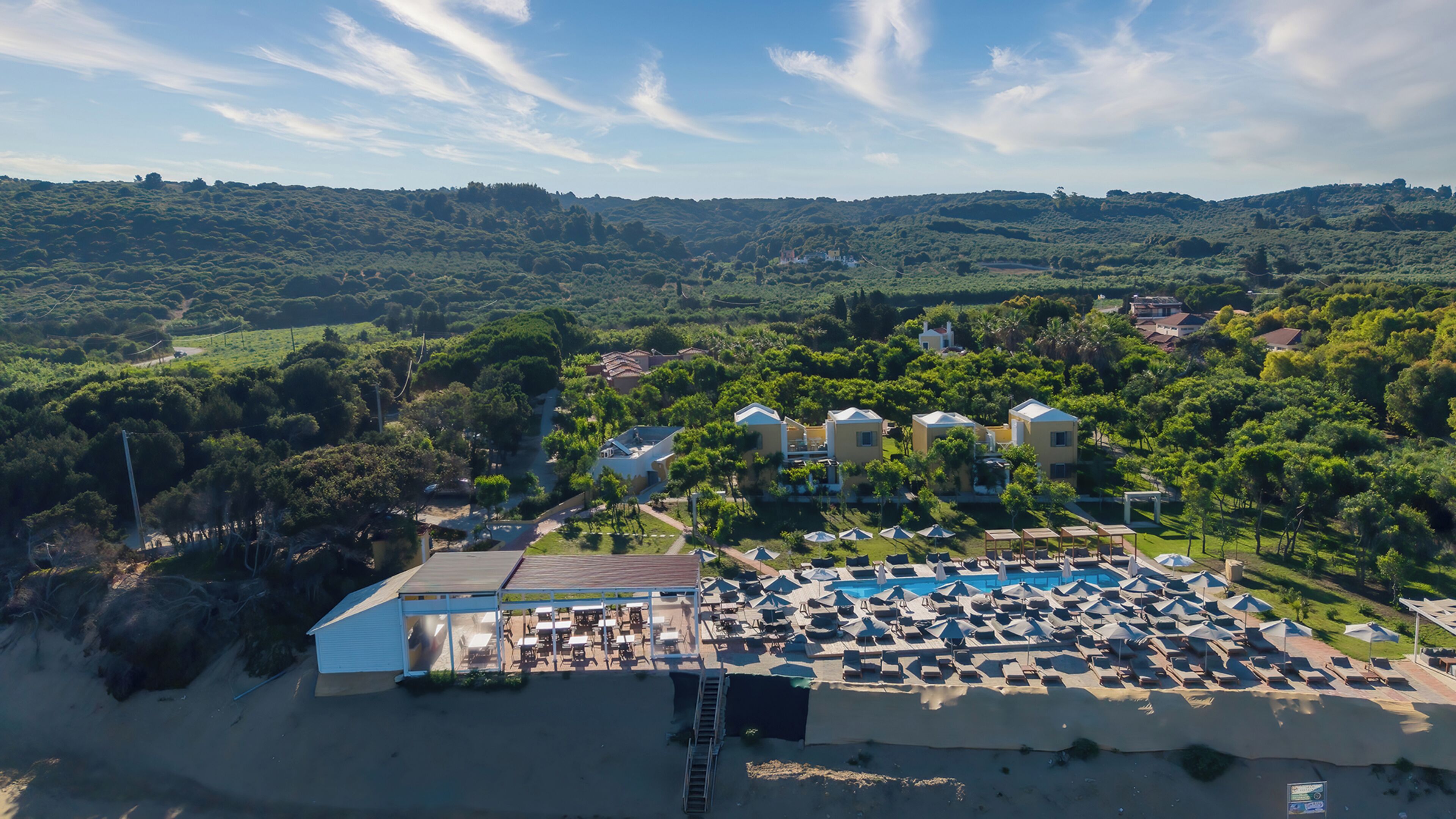 Vista aérea de un complejo turístico con piscina, tumbonas y vistas de las colinas circundantes.