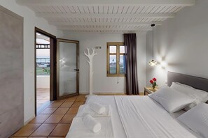 A hotel room with a bed, a view through a door, a window with curtains, and a pendant light.
