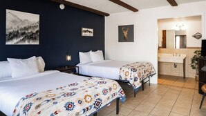 A hotel room with two beds, a nightstand with a lamp, a bathroom with a sink and mirror, and wall art. - The Alpine Motel (Collbran)
