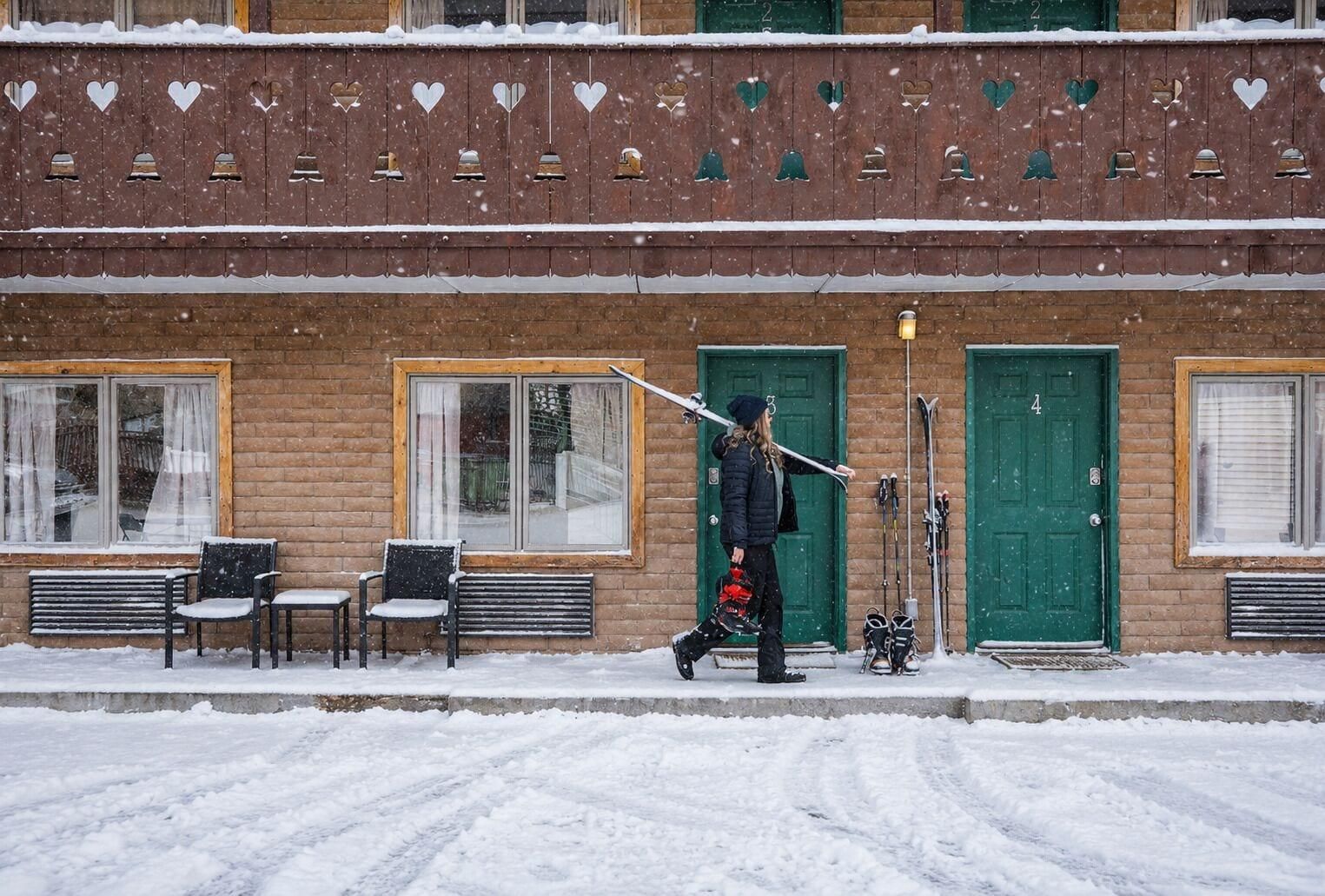 A building with a brick facade, multiple windows, and doors. There are benches outside and a person carrying skis.