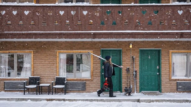A building with a brick facade, multiple windows, and doors. There are benches outside and a person carrying skis. - The Alpine Motel (Collbran)