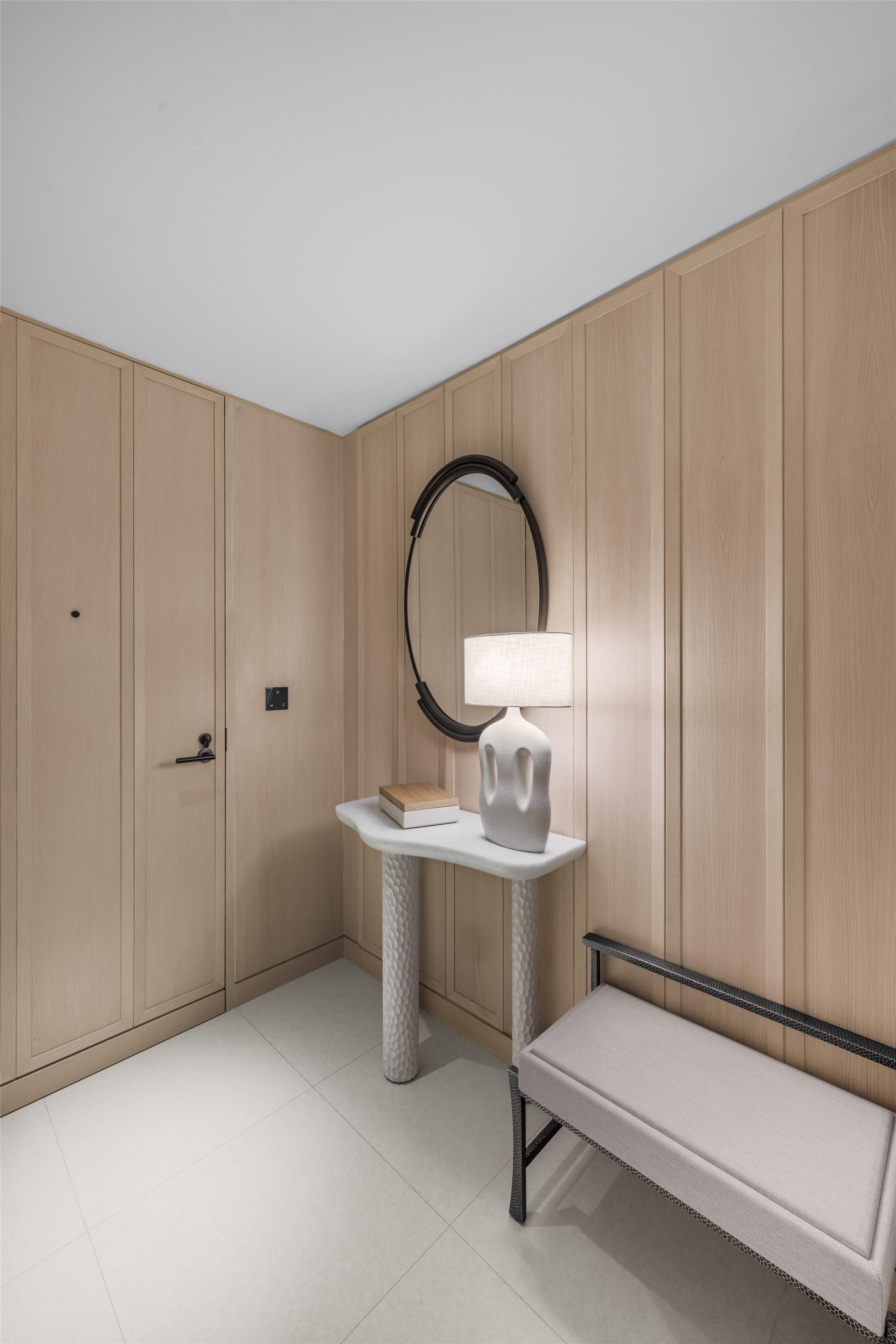 A room with a wooden wardrobe, a round mirror, a table with a lamp, and a bench.