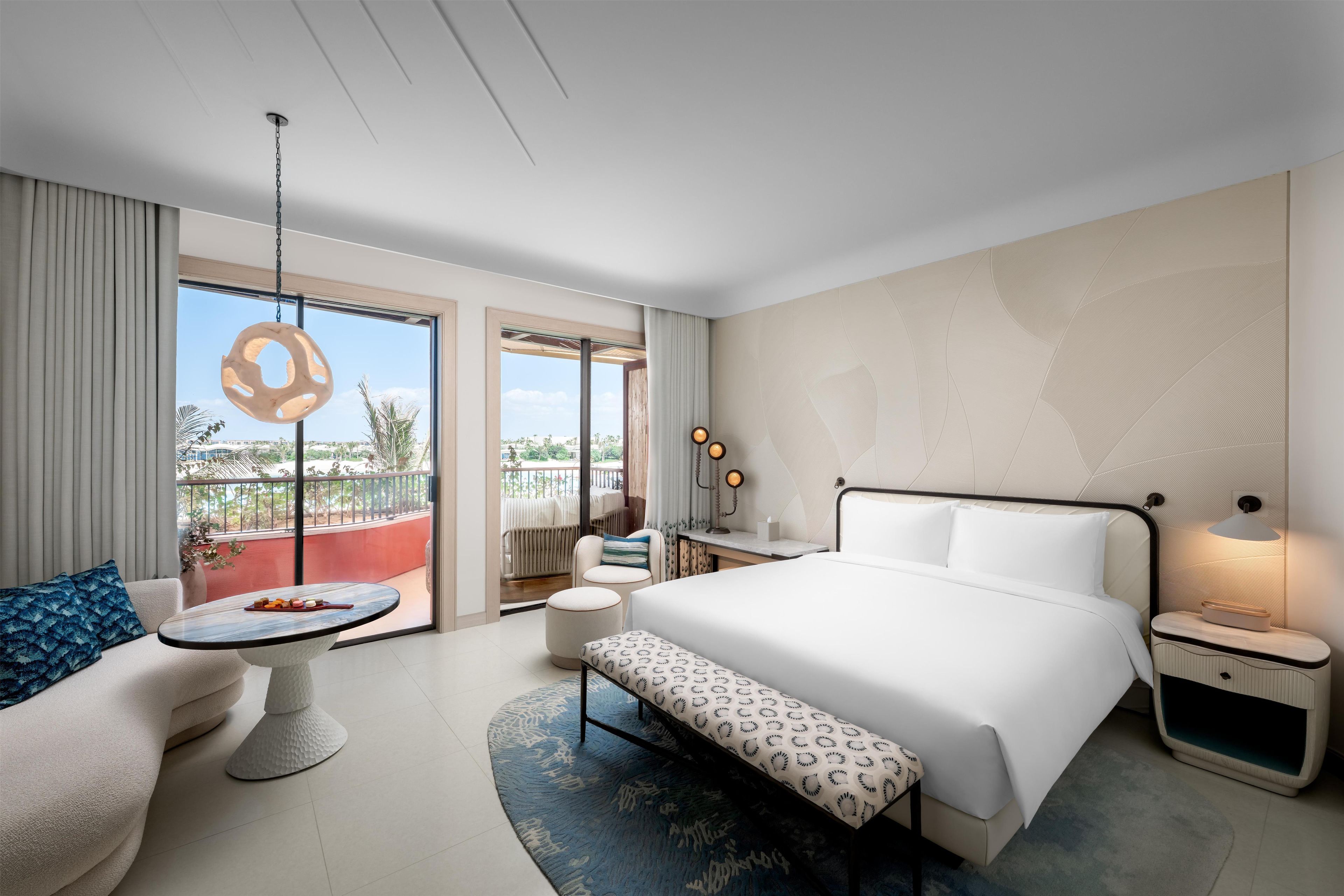 A modern hotel room with a large bed, a round table, a bench, a nightstand, and a view of the outdoors.